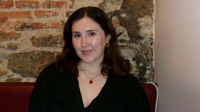 aimee sits in a red booth against a brick wall. she has shoulder length dark brown hair and wears a black v neck top with a gold necklace.
