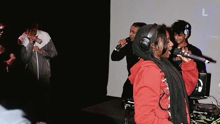 A lively scene of four people performing on stage. The focal person wears a red hoodie and headphones, singing into a microphone. Two others hold mics, with a DJ in the background. The atmosphere is energetic and engaging.