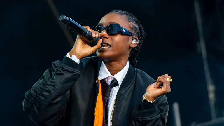 Little Simz wears a black jacket, white shirt, and sunglasses sings into a microphone on stage. The mood is energetic and vibrant, suggesting a lively performance.