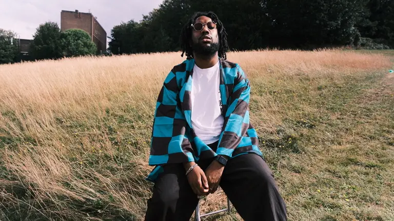 Tee Peters wears a blue checkered jacket and sunglasses sits on a stool in a grassy field, exuding a relaxed vibe. Trees and a building are in the background.