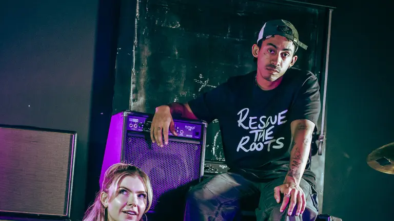 Jordan Stephens and Jodie Bryant sitting down in a. stage while wearing black t-shirt with Rescue the Roots 