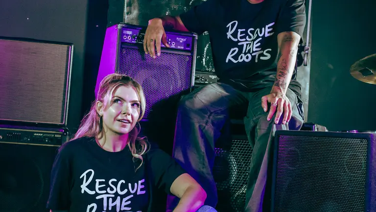 Jordan Stephens and Jodie Bryant sitting down in a. stage while wearing black t-shirt with Rescue the Roots 