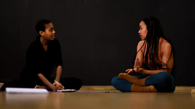 Two people sitting down in the floor, talking. One of them is taking notes in a a notepad.