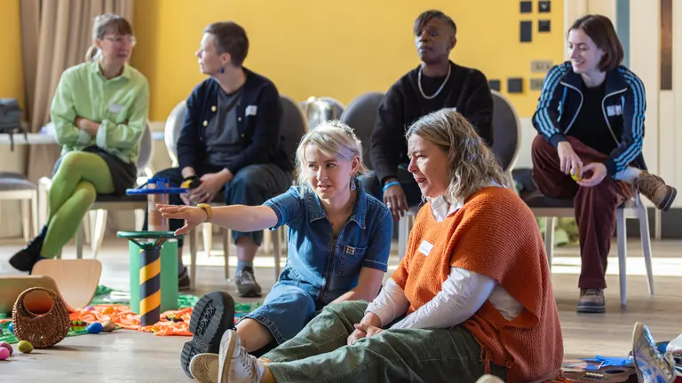 two adults sat on the floor, one is pointing to something off screen. 4 other adults sat behind them on chairs chatting to each other in front of a yellow wall