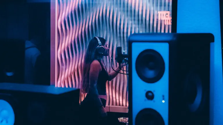 a young person in a vocal booth singing with headphones on, seen through the glass, where in the foreground you can see speakers and electrical equiptment. The light is a saturated blue
