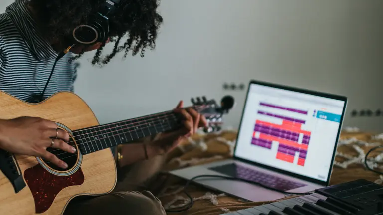 young person playing guitar with keyboard, headphone and a computer close