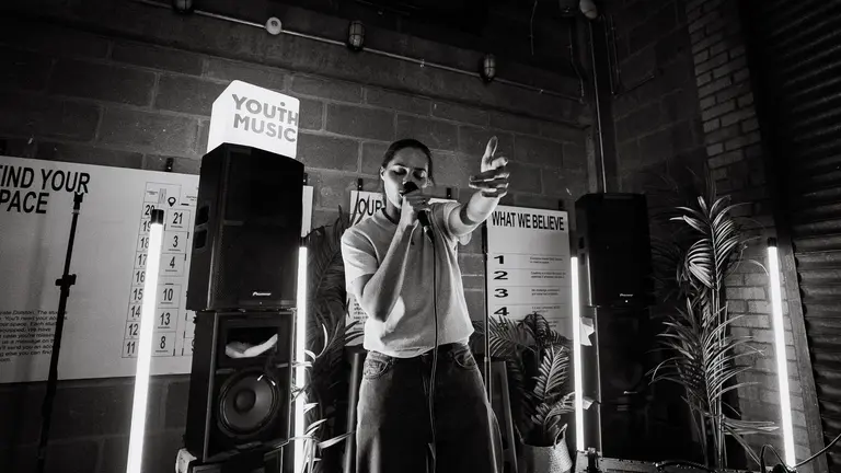 A young performer on stage, singing into the microphone with their arm reaching out to the audience. The image is in black and white and there is a lightbox with 'Youth Music' lit up