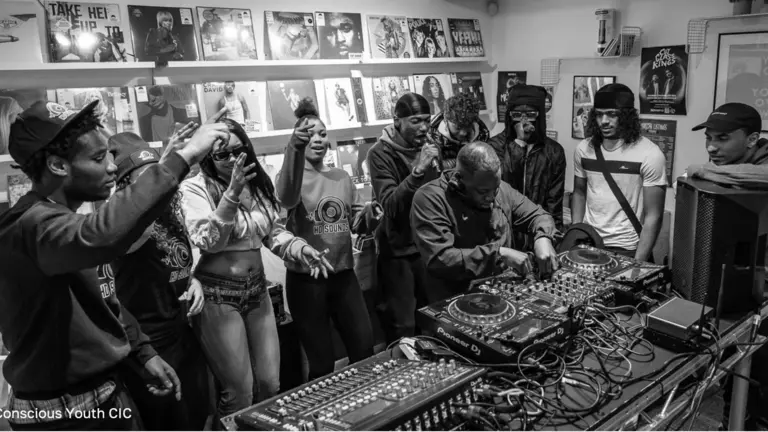 A group of young people with their hands up surrounding a DJ booth. The pic is in black and white. 