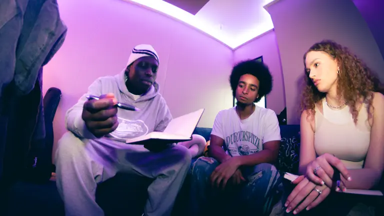 A fish eye lens image of three NextGen artists sat down. One man is holding a notebook and pen while the other two are looking at the notepad. 