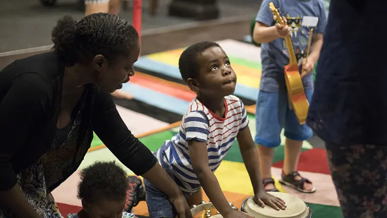 small boy in a play session with both hands on a drum standing up. Other children playing with instruments around him alongside adults