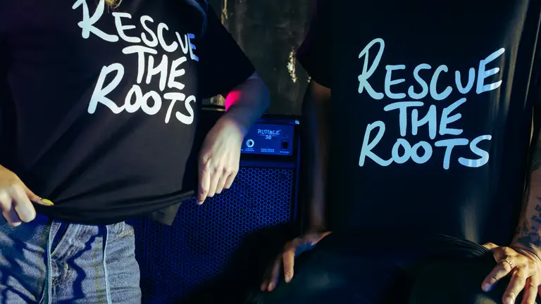Two people, with their heads not visible, with black shirts with white writing saying, 'Rescue The Roots'