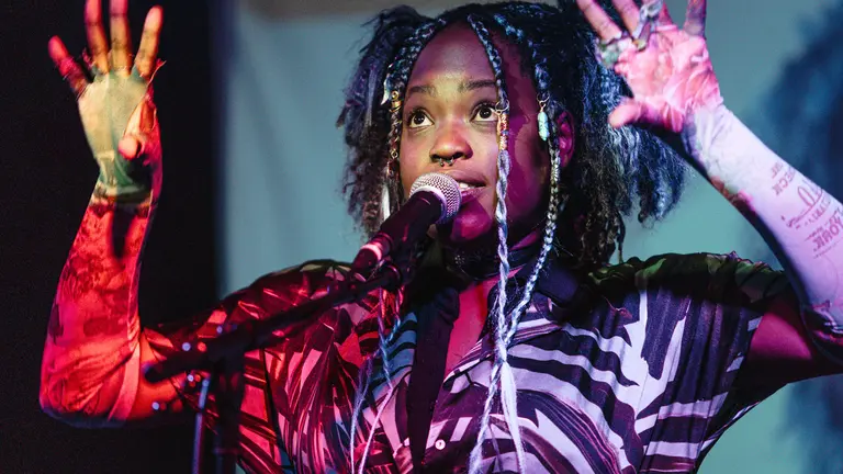 Woman on stage performing, with hands in the air, looking into the lights, in front of a microphone
