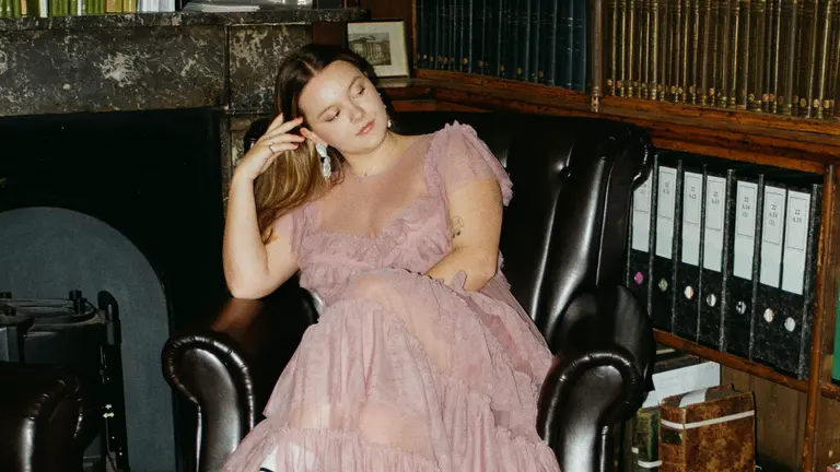 lauren wears a pink dress and sits in an armchair pensively