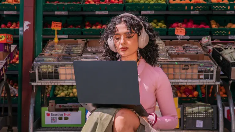 melika sits outside a grocer. she works on a laptop and wears headphones