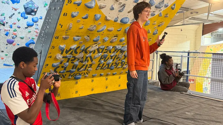 a man in a football shirt videos a woman in an orange jumper holding a microphone at a climbing wall