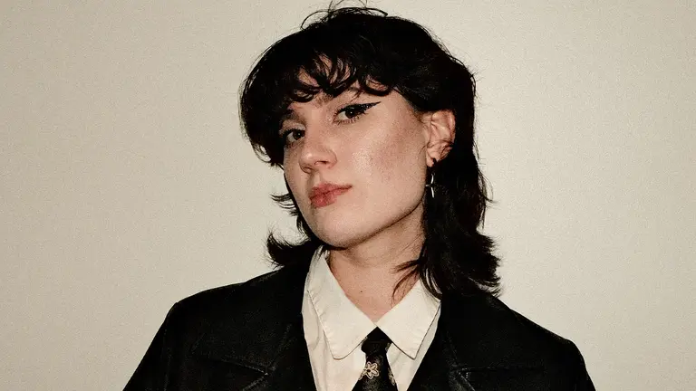 a woman with curly dark hair wears a shirt, blazer and tie