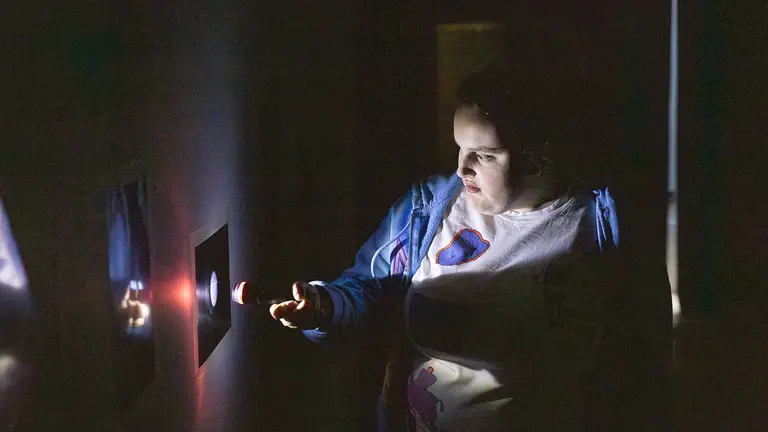 A person in a dimly lit space points a small flashlight or laser-like light toward an illuminated display on the wall, revealing glowing shapes in the darkness.