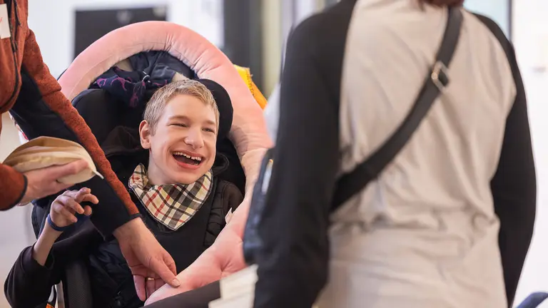 A person sits in a supportive wheelchair with a cushioned headrest, while two adults nearby interact with them—one reaching toward the chair and another standing facing them.
