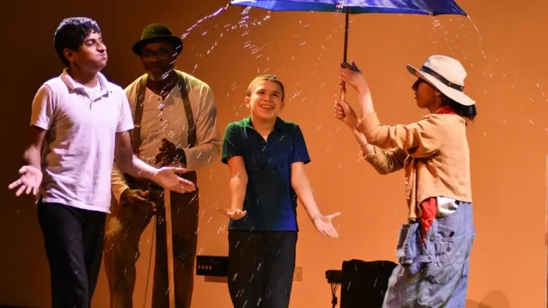 Four performers onstage take part in a playful scene where one holds a blue umbrella that splashes water outward, while the others stand with open arms reacting to the spray.