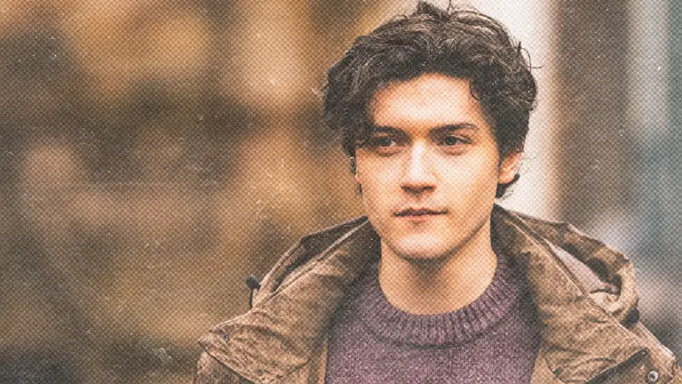 a young man with dark curly cropped hair wears a brown jacket and purple knitted jumper