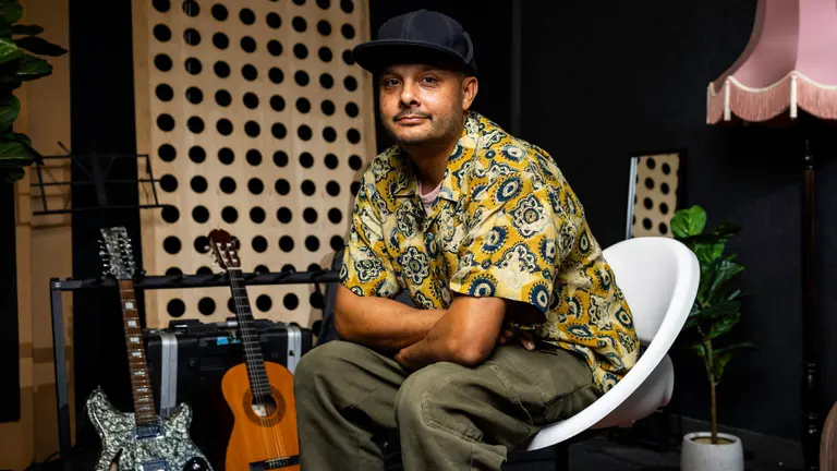 Adam sits on a white chair in a music studio, wearing a patterned shirt and cap. The room includes two guitars, acoustic treatment panels.