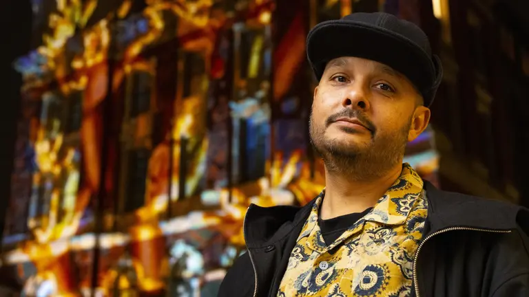 Adam stands outdoors at night wearing a patterned shirt and dark cap, with a brightly lit building behind them covered in colourful projected artwork.
