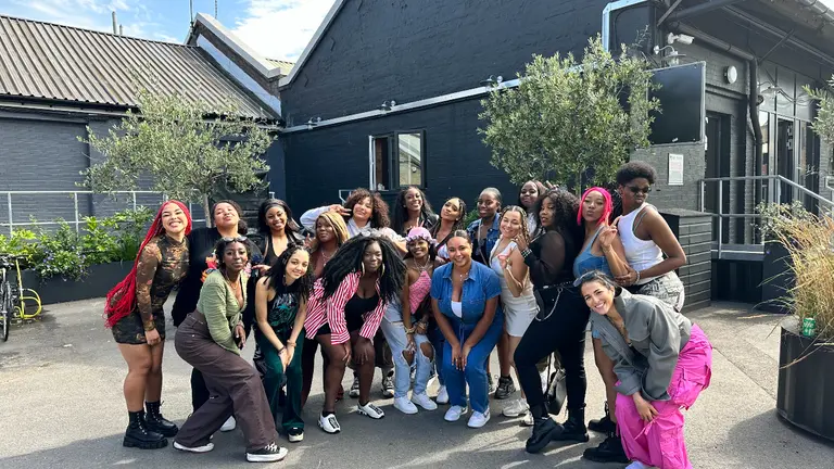 A large group of people from Girl Grind UK poses together outdoors in front of a dark grey building with a slanted roof. The group forms a close semi‑circle, with individuals standing or crouching in various dynamic poses.