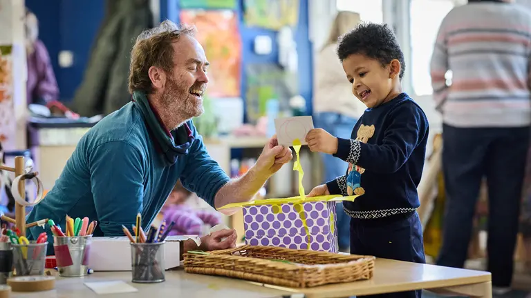 A child explores a ribbon inside a patterned box as an adult supports the activity. Art supplies and colourful classroom displays surround them, creating a playful early‑years learning environment.