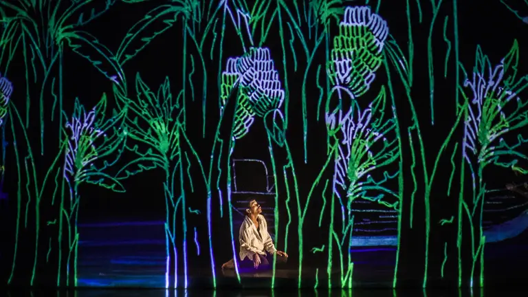 a young man on his knees on stage looks up at a large projection of trees on the back of the set