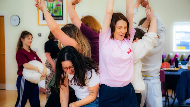 Early years practitioners in an active session, standing very close together, many with their hands in the air and smiling. 