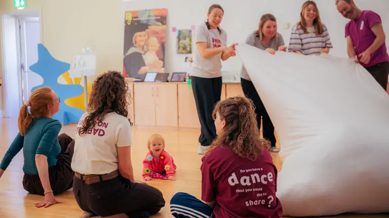 3 adult practitioners sat around a baby who is crawling and smiling, while 4 adults stand up holding a large pillow