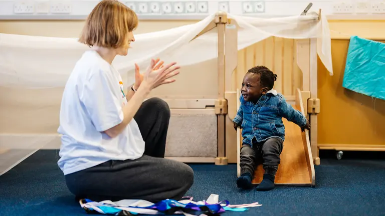 An early years child at the bottom of a slide indoors smiling, being clapped by a project leader