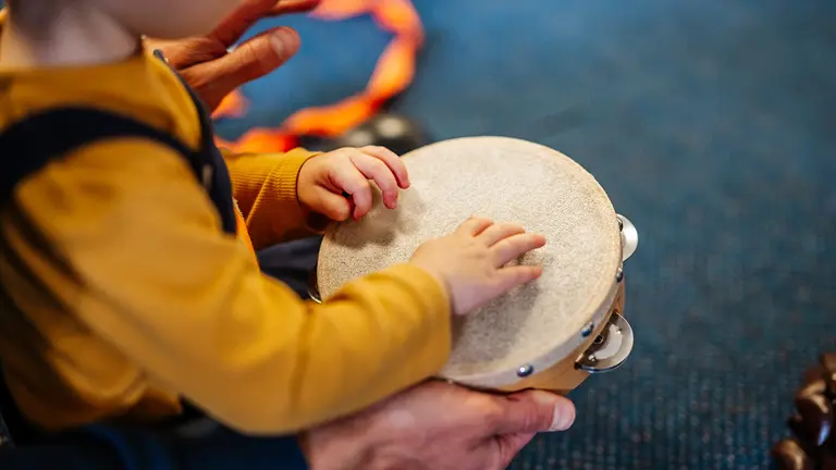 A picture of a child using a drum, it's wearing a yellow jumper and dungarees, you can't see the child's face