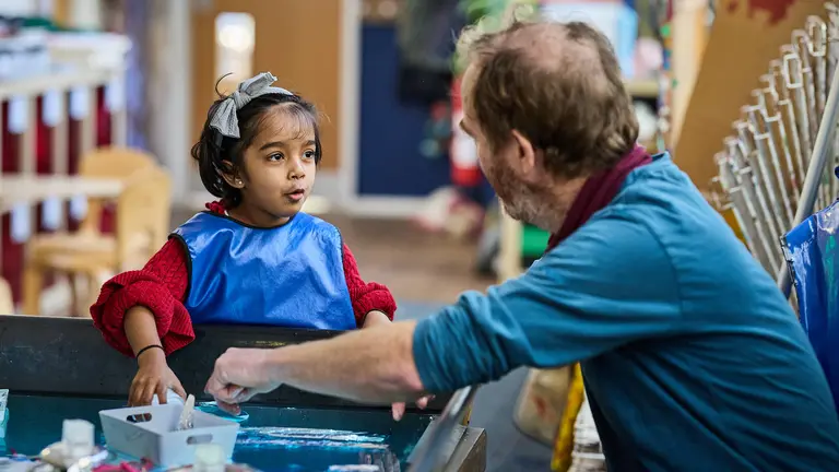 an early years child with a blue apron on playing with paints with a project leader joining in