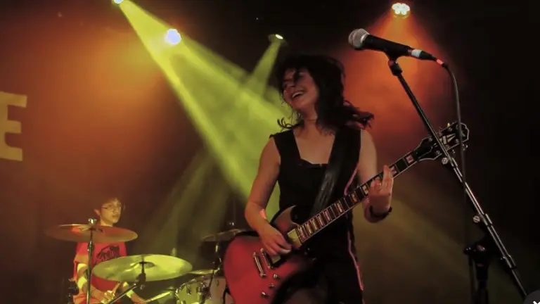 A young woman playing the electric guitar on stage, with a drummer behind and green/yellow lights