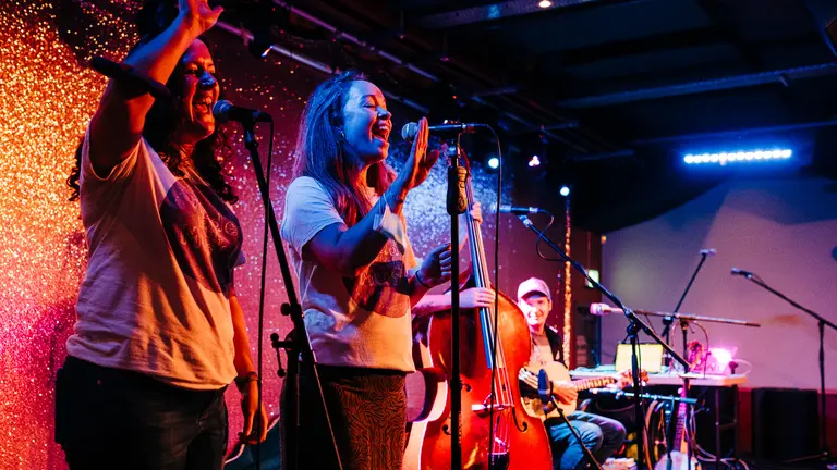 Made with Music on stage with two women singing with their arms in the air. Also a double bass on stage