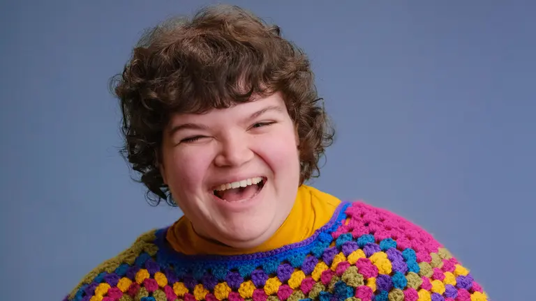 arwen. in a colourful jumper laughing and smiling at the camera with a blue/purple backdrop