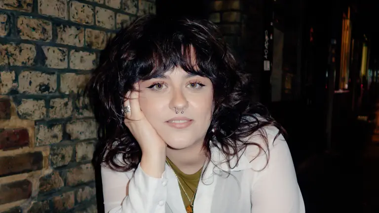 Headshot of Eve, resting her head on her hand, with a brick wall behind. 
