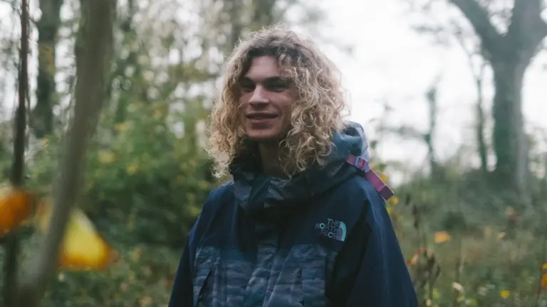 A person stood in a forest with shoulder length curly blonde hair and a rain jacket on