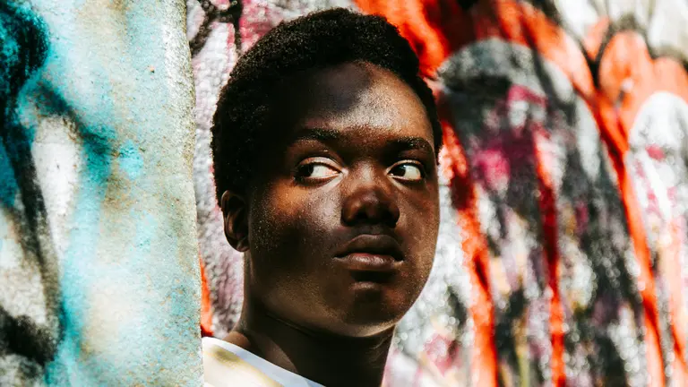 A man with his back to the wall looks to the right of camera in front of a colourful wall with the sun on his face