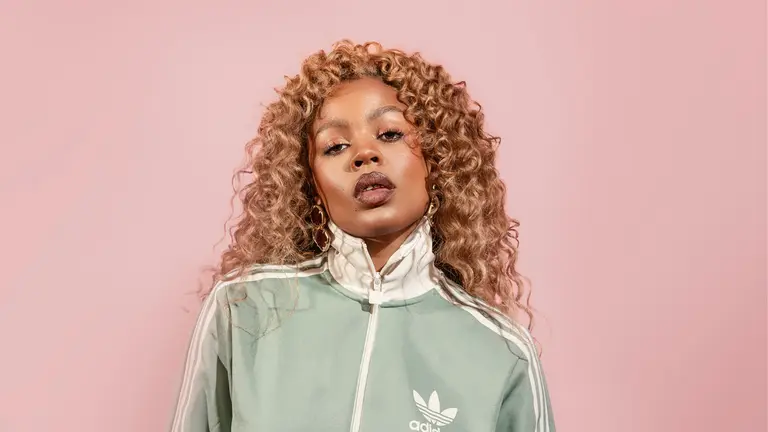 LAMAYA standing in front of a pink background, with a tracksuit on looking at the camera