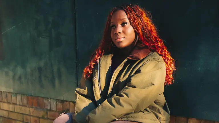 A girl sat outside with a jacket on, in the sun, looking off camera