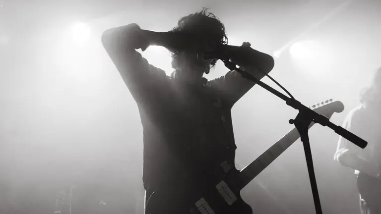 A sillhouette of the performer on stage with a microphone stand and guitar over them, with two arms behind their head