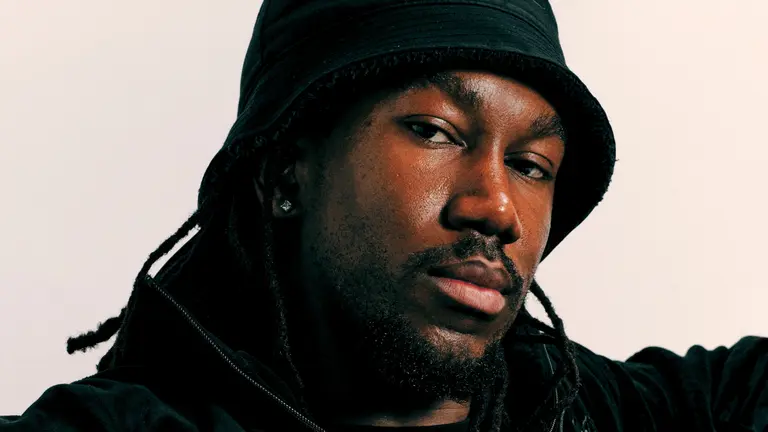 Mar!k headshot, with a bucket hat on, looking at the camera with a neutral expression, in front of a white/cream background
