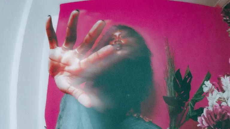 A woman putting her hand in front of her body with a very blurry filter, in front of a pink wall