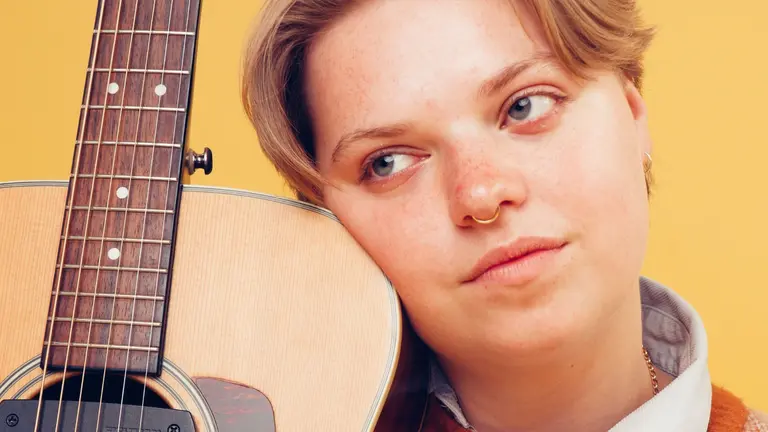 A person with their face against a guitar, looking into the distance, with a yellow background