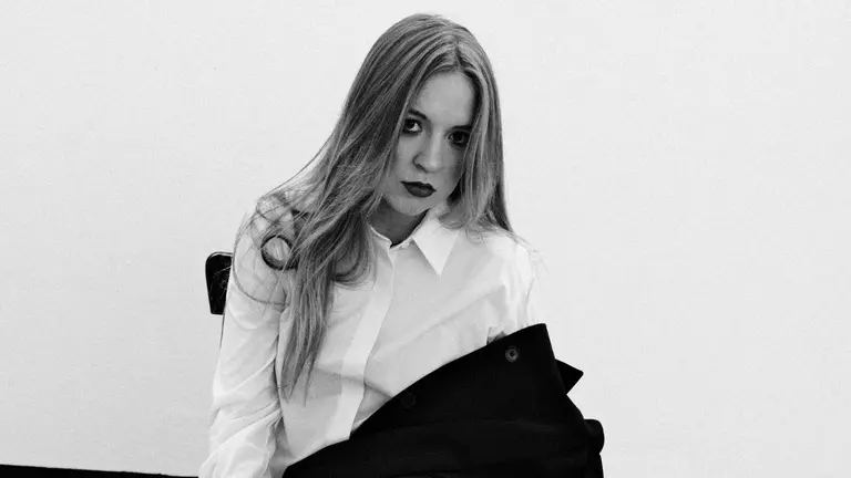 NEVATA sat in a white washed room, with a black jacket over their knees, looking intensely at the camera