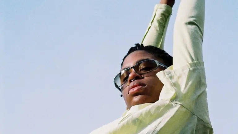 okcandice photographed from below, shades on looking at the camera, both arms above their head, with the blue sky as the background