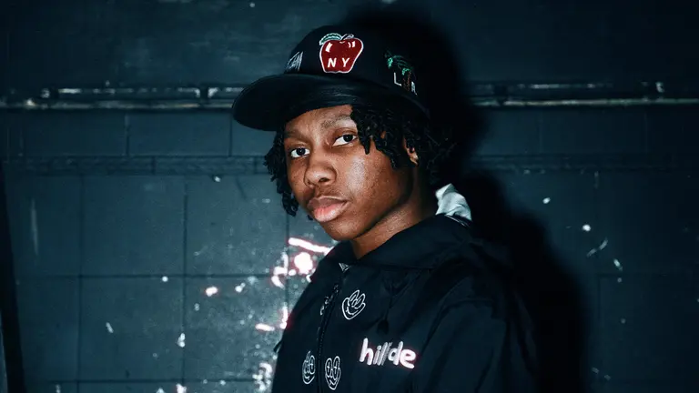 TBO with a cap on leaning against a wall and looking at the camera with a neutral expression