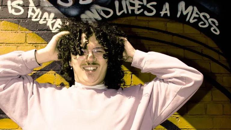 A man with a pale pink sweatshirt stands in front of a graffiti covered wall with both hands on his head